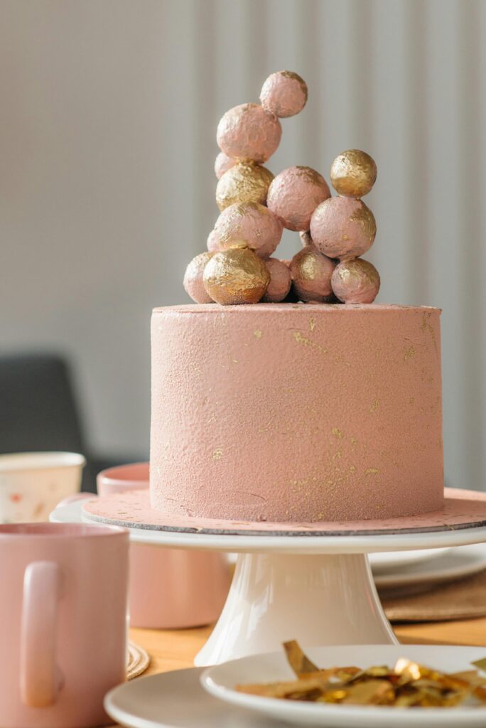 Pink cake topped with gold spheres, elegant design on a table with plates and mugs.
