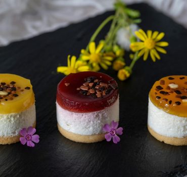 Delight in colorful mini fruit cheesecakes, beautifully garnished with flowers on a dark slate platter.