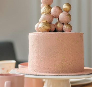 Pink cake topped with gold spheres, elegant design on a table with plates and mugs.